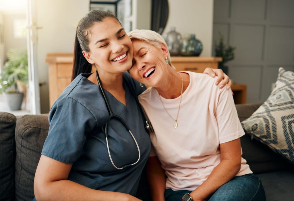Nurse and older senior woman sitting on couch hugging and smiling